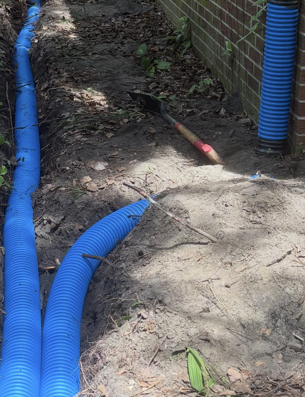 Two blue corrugated drainage collection lines set in an open trench beside a brick home