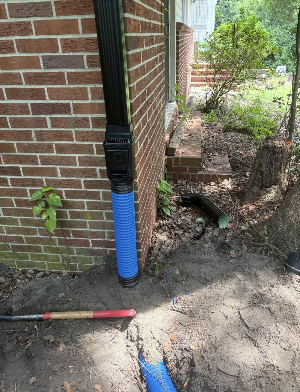Downspout connected into a buried corrugated drainage line at the corner of a brick home, with the backfill tamped around the connection