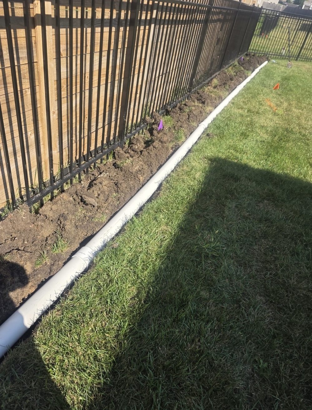 Solid PVC drain line set in a shallow trench along a fence in a Pleasant Hill, Missouri backyard, with 811 utility-locate flags placed before digging