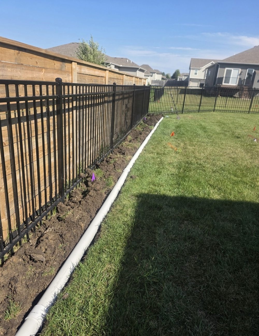 Long run of solid PVC drainage pipe laid along a backyard fence line in Pleasant Hill, Missouri, with utility-locate flags marking the route