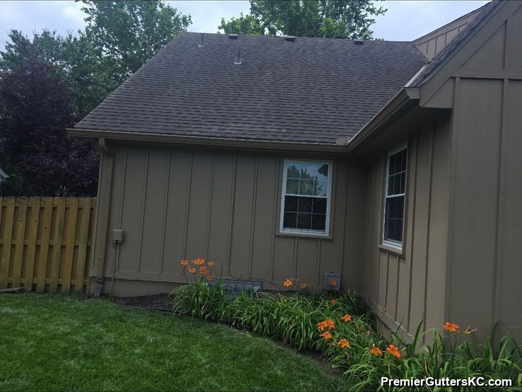 Tan board-and-batten home corner with seamless gutters along the roofline, bordered by an orange daylily bed and privacy fence in the Kansas City metro