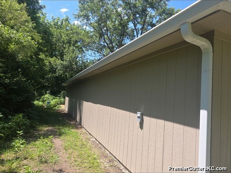 Side elevation of a tan ranch home showing a seamless gutter run and downspout carrying water away from the foundation toward a wooded side yard in the Kansas City metro