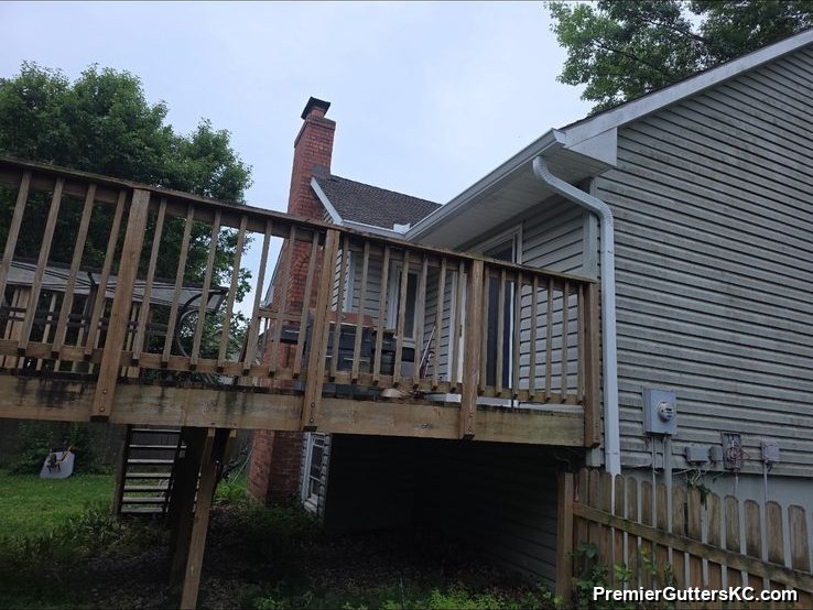 White downspout running down the side of a sage-green walkout ranch home past a raised wood deck and utility meters, draining away from the foundation in the Kansas City metro